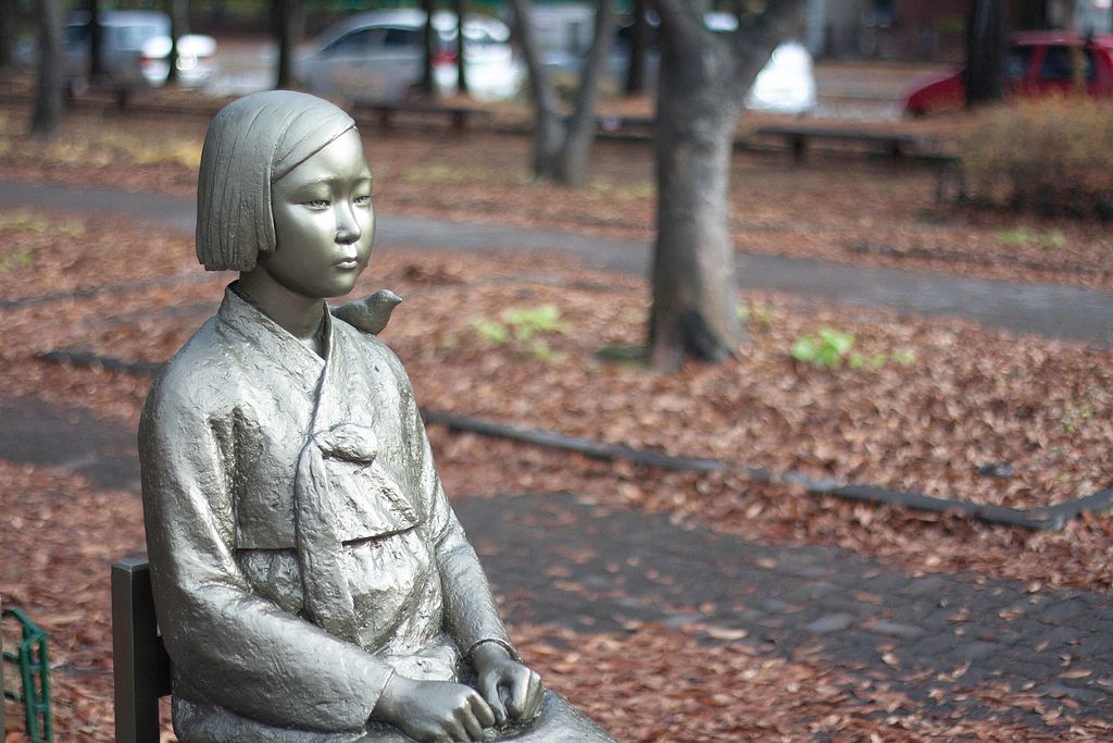 Statue of Peace. Sad looking Korean woman in traditional garb with clenched fists. Park-like background with tree trunks and leaves on ground. Autumn setting.(Photo By YunHo LEE - peace statue comfort woman statue (3), CC0)