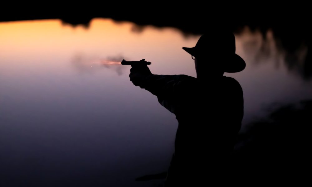 Silhouette of man in hat, shooting pistol over water.