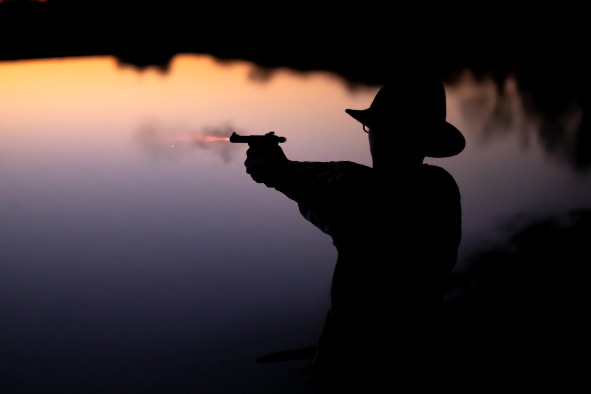 Silhouette of man in hat, shooting pistol over water.