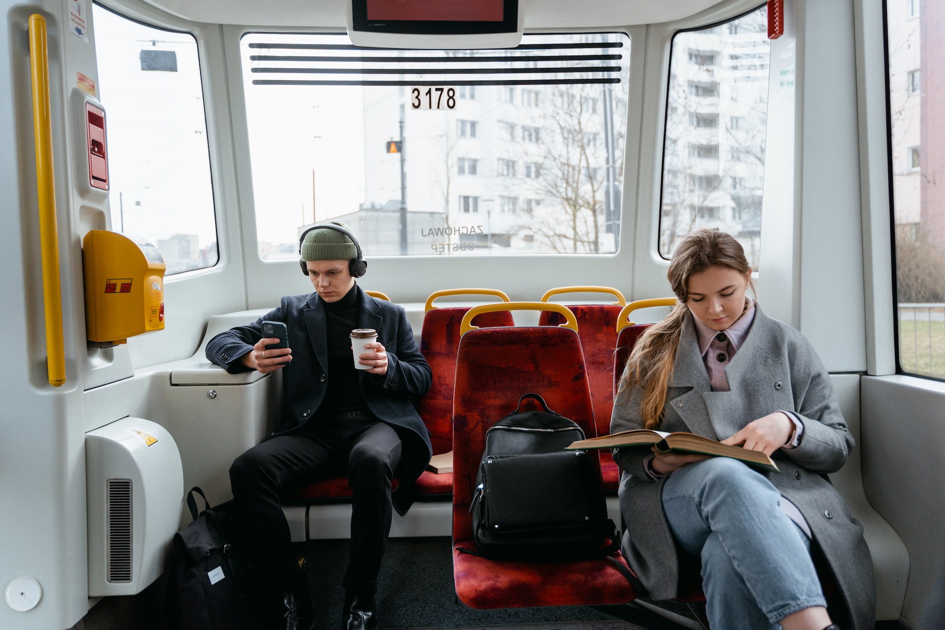 A Woman in Gray Coat Reading a Book while Sitting Near the Man Wearing Headphones while Holding His Mobile