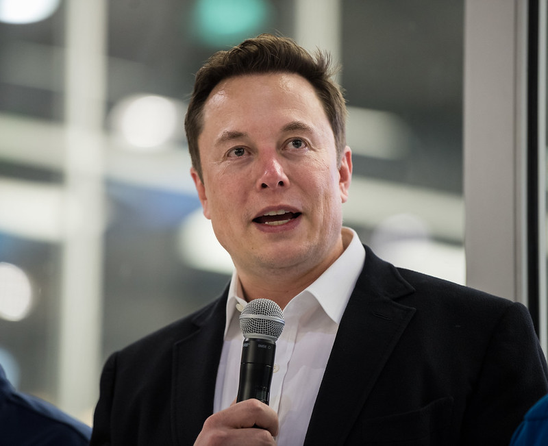 Elon Musk wearing black coat holding mic