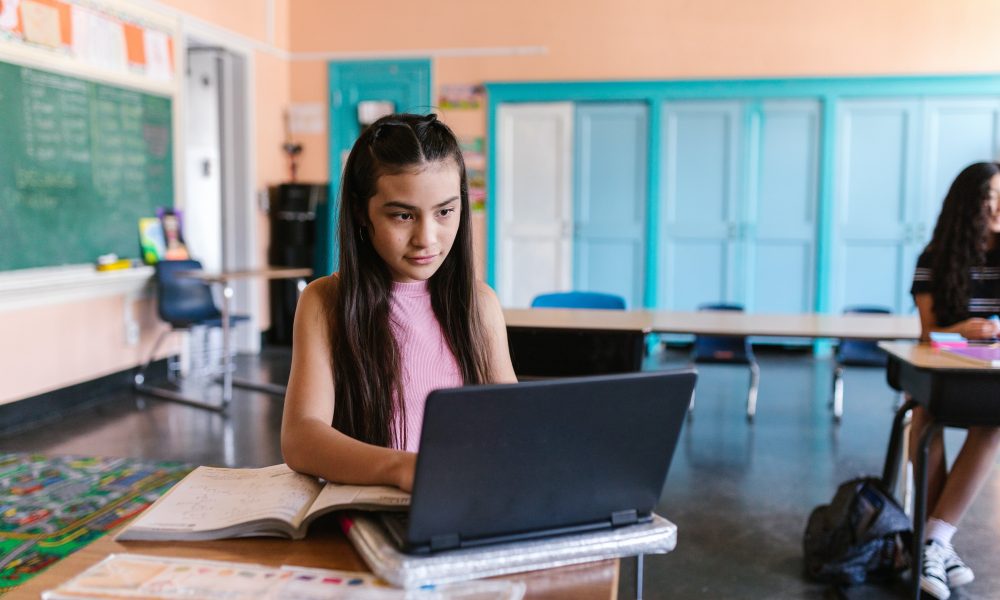 A Young Girl Typing on Her Laptop while Sitting on the Chair Inside the Classroom