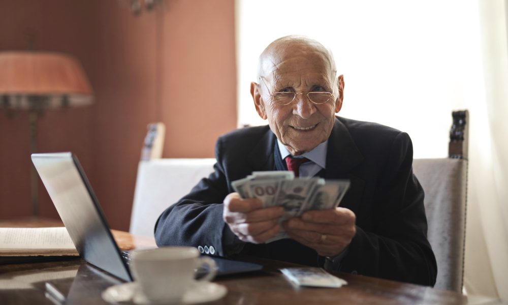 Confident senior businessman holding money in hands while sitting at table near laptop