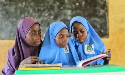 A Pair of Girls Wearing Blue Hijabs Reading a Book Beside a Young Woman Wearing Purple Hijab