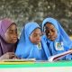 A Pair of Girls Wearing Blue Hijabs Reading a Book Beside a Young Woman Wearing Purple Hijab