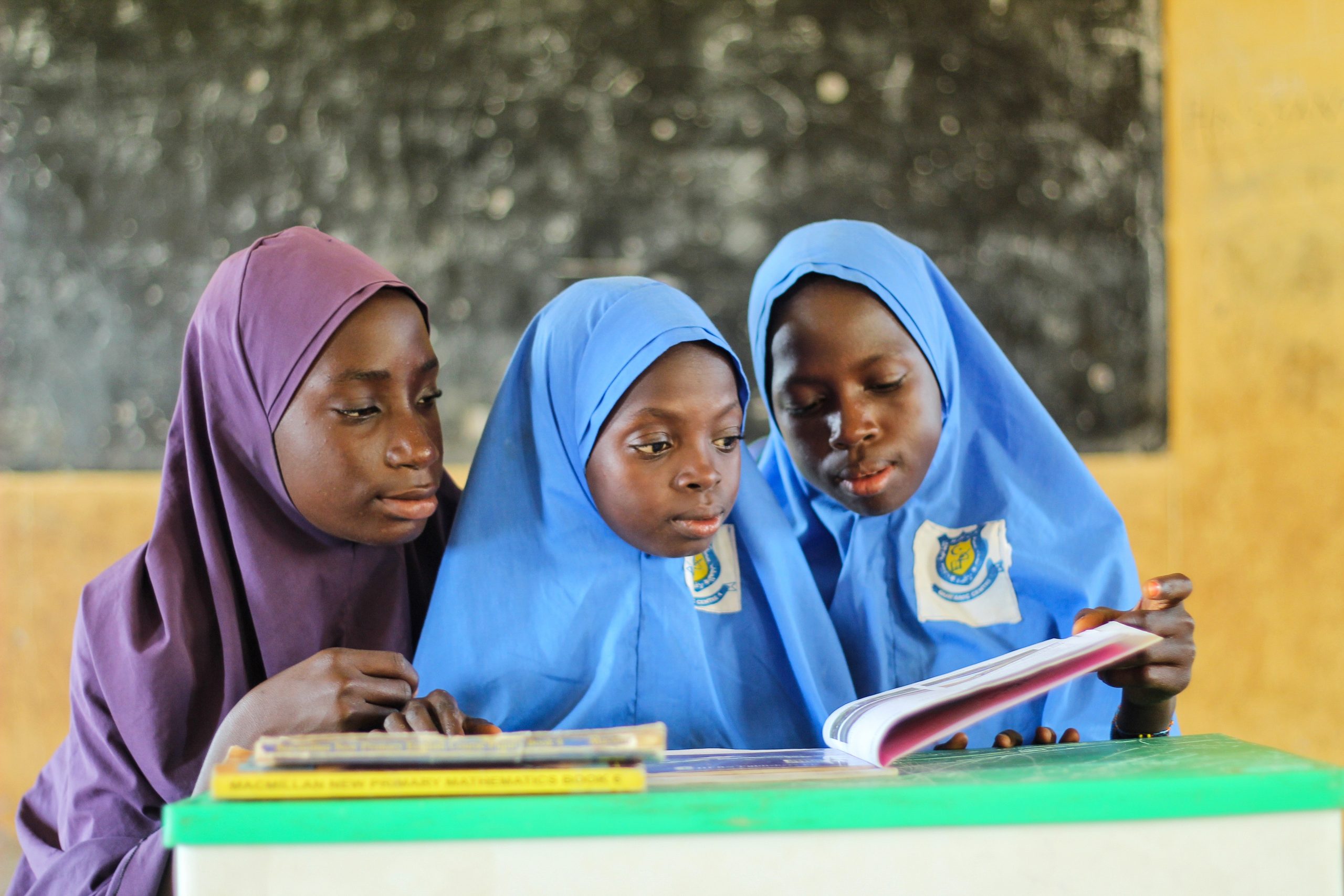 A Pair of Girls Wearing Blue Hijabs Reading a Book Beside a Young Woman Wearing Purple Hijab