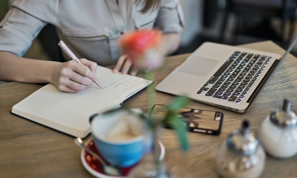 Person Writing On A Notebook Beside Macbook