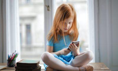 Calm small ginger girl sitting on table and using smartphone in light living room