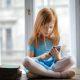 Calm small ginger girl sitting on table and using smartphone in light living room