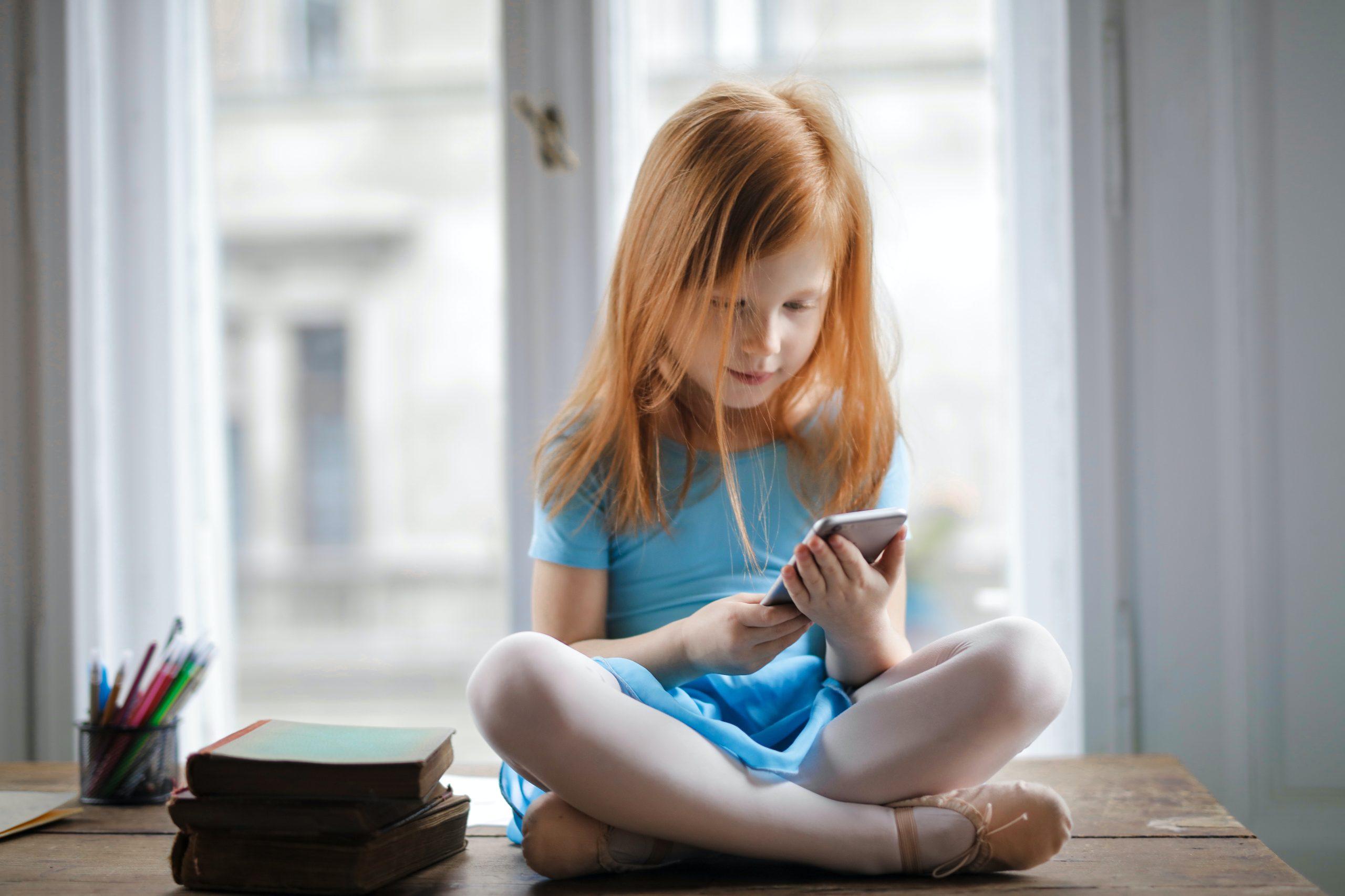 Calm small ginger girl sitting on table and using smartphone in light living room