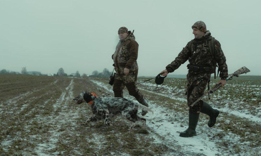 Two Men in Camouflage Walking in the Field with a Dog