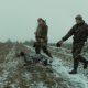 Two Men in Camouflage Walking in the Field with a Dog