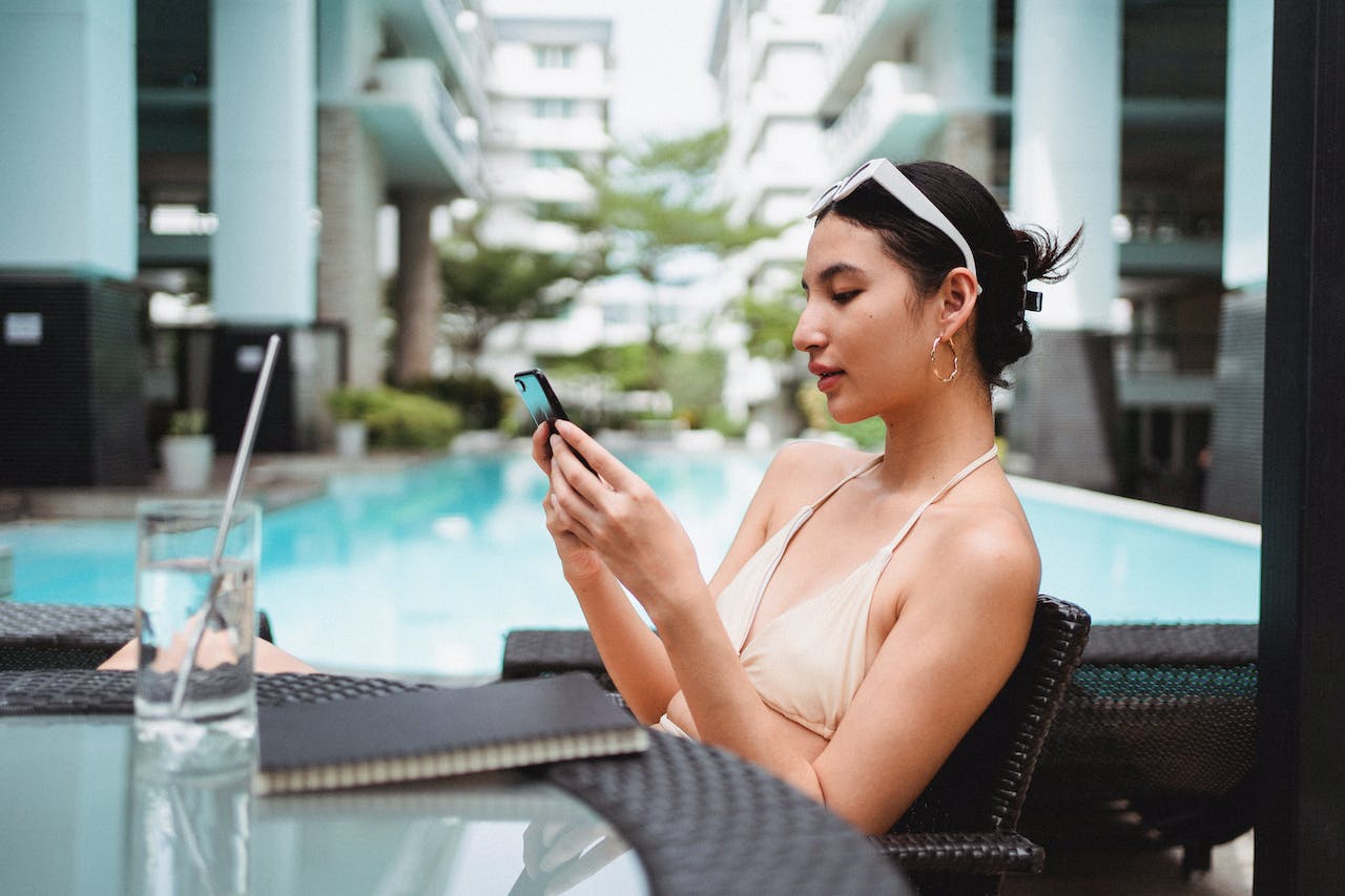 Young ethnic woman messaging on smartphone while chilling in cafe near outdoor pool