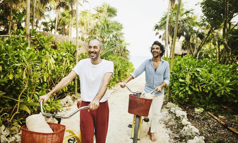 Medium wide shot of laughing gay couple walking bikes on path from beach at tropical resort