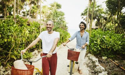 Medium wide shot of laughing gay couple walking bikes on path from beach at tropical resort