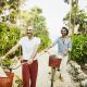 Medium wide shot of laughing gay couple walking bikes on path from beach at tropical resort